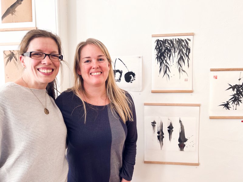 Jo and Meah stand in front of a wall of their ink brush paintings