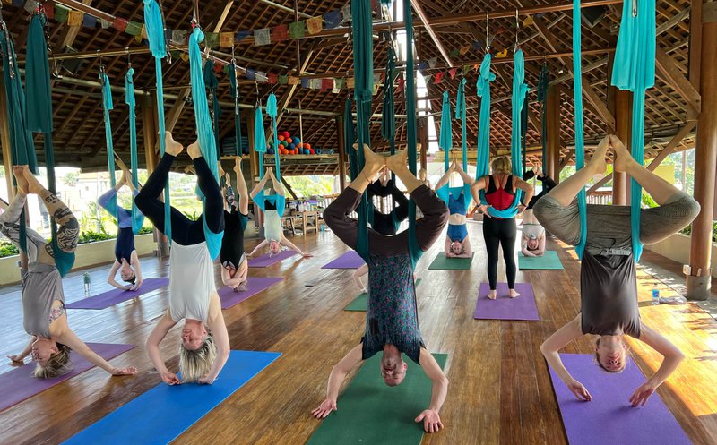 12 people are hanging upside down in aerial hammocks in a traditional Balinese bamboo building