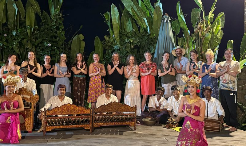 In the foreground we see two female Balinese dancers in traditional dress, behind them we see six male musicians with ornately carved wooden instruments. In the back row are the 14 retreat guests dressed in colourful outfits.